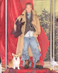 A person stands confidently against a vivid backdrop with rich textures and patterns. They wear a layered ensemble featuring an oversized coat and strikingly unique jeans. At their feet is a cheerful corgi, adding a playful touch. Nearby, a pigeon observes the scene. The overall composition is bold, blending fashion with whimsical elements.
