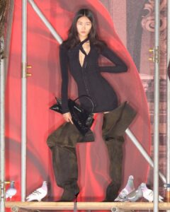 A person stands confidently in a fitted, dark, long-sleeved outfit with a plunging neckline, complemented by oversized suede boots. They hold a sleek black bag with a bold design. The setting is framed by metal scaffolding against a red backdrop, adorned with pigeons casually perched along the bottom, adding an urban touch to the scene.