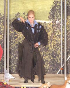 A striking figure poses confidently in an intricate ensemble, featuring a black textured coat over a blue collared shirt. The backdrop displays lush greenery, while a pigeon perches beside and a dog sits nearby. The setting blends elements of nature with an urban, artistic sensibility. The person exudes elegance, adorned with bold makeup and a statement necklace.