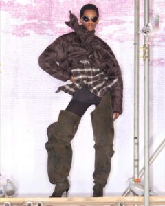 A model exudes confidence wearing an avant-garde ensemble featuring a voluminous, puffy brown jacket and a patterned flannel shirt. The look is completed with over-the-knee, high-heeled, slouchy boots. Sunglasses add a chic touch. On either side, pigeons are perched on metal pipes against a textured pink and white background, creating an urban aesthetic.