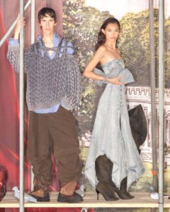 Two individuals stand on a wooden platform. The person on the left has an avant-garde fashion style with a layered gray knit sweater over a striped shirt and brown pants. The other individual wears a striking light blue strapless dress paired with high boots. They are set against a backdrop featuring greenery and architecture, with small decorative birds nearby.
