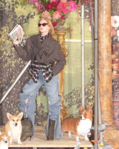 A fashionable individual with long hair and sunglasses stands confidently in layered outfits, holding magazines. The scene is adorned with lavish floral arrangements in the background. Ripped jeans and unique footwear complement the look. Around them are two small dogs, one real and one a statue, with a decorative, artistic setting featuring pillars and rich textures.