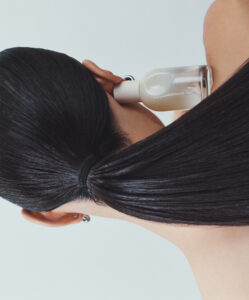 A person with long, smooth black hair is tied in a low ponytail, facing away. They hold a translucent bottle with a white cap near their face, suggesting a moment of contemplation or use. The background is light, and the person wears a small hoop earring. The scene conveys a sense of elegance and simplicity in personal care.