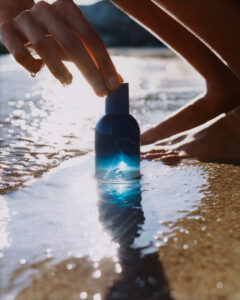 A hand reaches over a small blue bottle sitting on wet sand, reflecting sunlight. Droplets of water cling to the fingers, suggesting a beach setting. The sun casts bright highlights across the scene, creating a shimmering effect on the surface. The scene evokes a sense of freshness and relaxation, typical of coastal environments.