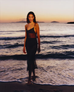 A woman stands on a beach during sunset, with the sky transitioning from orange to purple hues. She wears a sheer, black dress and a delicate necklace, her wet hair suggesting a recent swim. The calm waves gently lap at her feet, while distant islands outline the horizon, creating a serene and peaceful atmosphere.