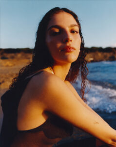 A woman with wet hair gazes thoughtfully. Her skin glows under the warm, golden light, casting gentle shadows. She wears a sleeveless black top, and her expression is serene. The backdrop captures a serene coastal landscape with gentle waves approaching a rugged shoreline, while the sky transitions from daylight to dusk, enhancing the tranquil scene.