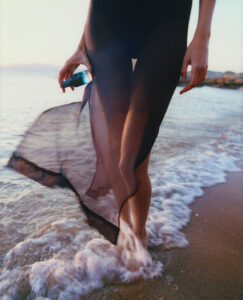 A person, partially visible, stands at the edge of the sea, where gentle waves meet the sandy shore. Their attire is sheer and flowing, with water lapping around bare feet, creating a serene and elegant atmosphere. The backdrop of a tranquil sunset and rippling water suggests a peaceful moment. They hold a small, rounded blue object.