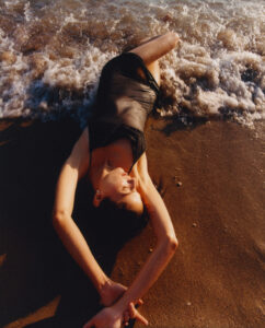 A person in a black outfit lies on wet sand near the water's edge, with waves gently lapping at their body. Their arms are elegantly extended above their head, fingers interlocked. Sunlight casts a warm glow, highlighting their relaxed expression. The dynamic contrast between the soft sand and foamy waves creates a serene and contemplative atmosphere.