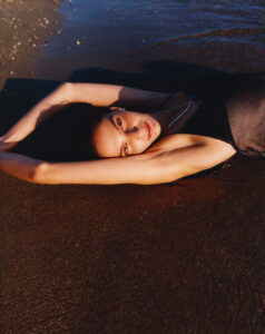 A person stretches out on wet sand by the water's edge, wearing a dark outfit. With arms gracefully extended above the head, they gaze directly upward, bathed in warm sunlight that highlights their features. The contrast between the textured sand and shimmering water creates a serene and captivating atmosphere, evoking a sense of tranquility and connection with nature.