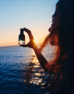 A figure stands beside the sea at sunset, holding a small glass bottle toward the fading light. Warm golden rays filter through their fingers as the horizon glows in soft orange and blue tones. Wind catches loose strands of hair, adding movement and intimacy. Reflections shimmer across the water, creating a dreamy atmosphere of calm, wonder, and quiet contemplation.