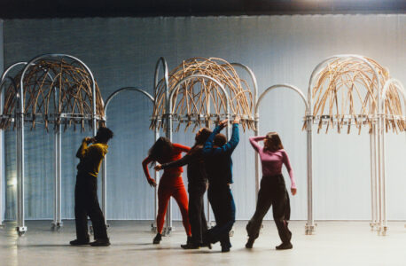 Five people engage in expressive movement beneath intricate metallic arches with woven branch structures. The individuals wear colorful, contrasting outfits in a spacious setting, marked by a soft, diffused light. The scene has an artistic, performance-like quality, capturing a blend of motion, creativity, and interaction against a minimalist, modern backdrop, highlighting a dynamic fusion of human expression and architectural elements.