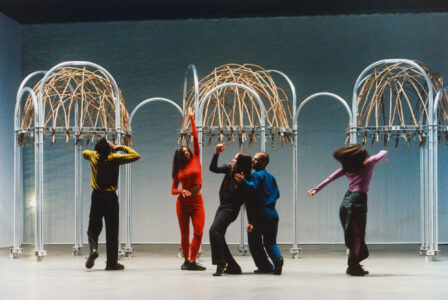 Five individuals engage in expressive movement beneath tall, arching structures with woven elements. The figures wear colorful, contrasting outfits—yellow, red, black, blue, and purple—enhancing the dynamic scene. Their poses are varied, suggesting a sense of rhythm and creativity. The background is neutral, accentuating the architectural elements and the lively motion of the performers.
