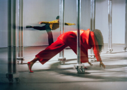 Two individuals engage in expressive dance amidst a series of vertical metal poles on wheels. One person, clad in vibrant red, is in an athletic pose, leaning forward with extended limbs. The other, dressed in mustard yellow, balances gracefully with one leg raised. The scene captures a sense of fluidity, movement, and artistic expression in a minimalist setting.