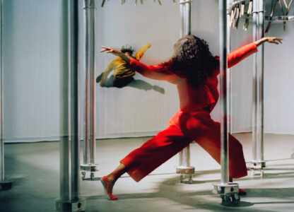 A dynamic scene shows an individual in vibrant red attire striking an expressive pose with outstretched arms amidst metallic poles on wheels, suggesting a sense of motion. In the background, another person in mid-air appears to be captured in a leap or dance. The setting suggests an artistic, possibly performance-based environment, creating an energetic and dramatic mood.
