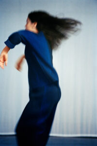 A person with long hair moves gracefully, wearing a dark blue outfit. The motion captures dynamic energy, with hair and limbs slightly blurred against a softly neutral background. The sense of movement suggests a dance or a spontaneous expression, creating a vibrant, fluid impression while focusing on the elegance and freedom of the action.