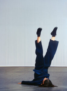 A person wearing a dark blue outfit performs a headstand on a dark surface. Their body is inverted, with hands on the ground and legs pointed upward. The background is a plain, light-colored backdrop. Their black shoes contrast with their attire, and the scene conveys a sense of balance and poise within a minimalist setting.