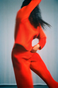 A dynamic figure clad in vibrant red appears in mid-motion, with a blur suggesting dance or energetic movement. The long, wavy hair flows freely in sync with the graceful pose. The soft background contrasts with the bold hue, emphasizing the sense of motion and rhythm, creating an aura of fluidity and artistic expression.
