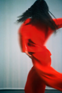 A dynamic scene features an individual with long, flowing hair moving swiftly, creating a sense of motion and urgency. The person is wearing a vibrant red outfit, which adds energy and contrast against the soft, blurred background. The movement and colors combine to evoke a sense of fluidity, creativity, and artistic expression.