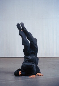 A person dressed in all-black attire, including a hood and mask, performs a headstand on a smooth, gray surface. Their legs are pointed upwards, demonstrating balance and flexibility. The background features a plain, light gray wall, creating a minimalist and focused atmosphere. Their hands are positioned on the ground for support, highlighting physical skill.