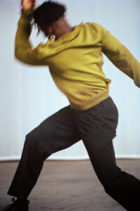 A dynamic scene captures a figure in motion, dressed in a vibrant olive-green top and dark pants. The movement is fluid, suggesting expressive dance or athleticism. The blurred motion conveys energy and spontaneity. The backdrop is simple, with a neutral tone, allowing focus on the subject’s gesture and form as they stride forward with intensity.