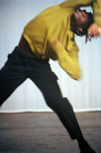 A person appears to be caught mid-movement, suggesting dynamic motion. They are wearing a yellow sweater and dark pants, with their body leaning sideways, creating a sense of fluidity and energy. The background is neutral, enhancing the focus on their motion. The captured moment conveys a sense of dance or expressive movement, embodying freedom.