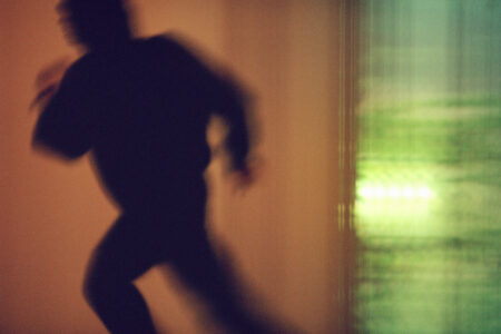 A shadowy figure appears in motion, rushing across a warmly lit space. The background blends soft orange and green hues, creating an abstract and dynamic scene. The silhouette's hurried posture suggests urgency, while the blurred lines and colors evoke a sense of fleeting movement, contrasting the comforting, muted tones surrounding the energetic subject.