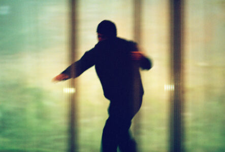 A blurred figure in motion, possibly running or sliding, is set against a soft, abstract background of muted green and yellow hues. The scene conveys a dynamic, almost dreamlike atmosphere, with the figure's outline suggesting purposeful movement. The combination of colors and distortion adds a mysterious, almost ethereal quality to the overall composition.