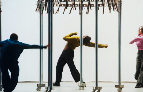 Three individuals are dancing energetically within a metal structure on wheels. One wears yellow, striking a dynamic pose, another in blue leans forward, and the third in pink moves gracefully. The background is minimal, emphasizing the performers' movements. The arrangement of the poles adds a geometric element, creating a sense of rhythm and motion.