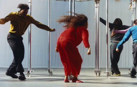 Four people are energetically dancing on a smooth, light-colored floor. Each wears vibrant attire: one in a yellow top, another in a striking red outfit, and others in blue and black. The scene captures dynamic movement, with hair flowing and limbs outstretched. Metal poles with wheels are arranged in the background, adding an industrial touch to the lively performance.