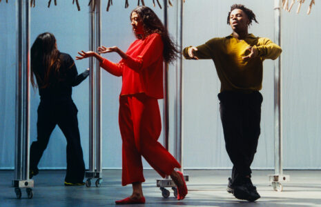 Three individuals are engaged in artistic movement within an industrial setting. The one in front, wearing a bright red outfit, appears to be in mid-step, exuding energy and fluidity. Another person in a mustard top and dark pants moves gracefully, while the third, dressed in black, faces away. Metal structures on wheels form the background, casting soft shadows.