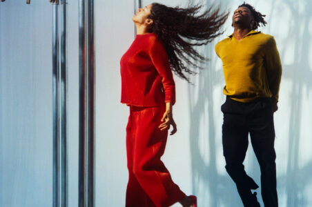 Two individuals are captured mid-movement. The person on the left wears a flowing red outfit, hair dramatically splayed. The right individual sports a mustard-yellow top and black pants, appearing poised and energized. The background is a smooth, light surface with vertical metallic elements, casting shadows. The scene conveys a sense of dynamic motion and vibrant expression.