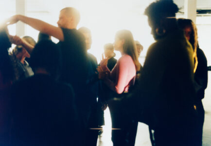 A dimly lit room with a gathering of people, some engaged in conversation. The atmosphere is casual, with individuals dressed in a variety of outfits, including a noticeable bright pink top. The setting suggests an informal social event or gathering, with soft, ambient lighting creating a warm, blurred background, adding a sense of intimacy and energy.