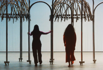 Two figures stand under structures resembling arches made from curved metal poles topped with intertwined branches. The ground is smooth and reflective, adding to the serene atmosphere. Both figures face away, one with outstretched arms touching the metal, the other walking forward. A soft, diffused light surrounds them, casting gentle shadows on the ground.