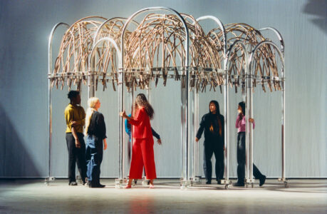 Six individuals interact with a modern, art-like structure composed of metal arches and intertwined wooden elements. The scene is set against a minimalist backdrop with soft, even lighting. Each person is uniquely dressed, suggesting a creative or artistic environment. The atmosphere is serene, emphasizing the contemplative nature of the setting. The focus is on the dynamic between people and installation.