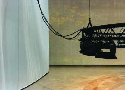 Stage setup with a curved white curtain on the left, casting soft shadows. On the right, a suspended black truss system hangs from a cable, fitted with equipment, possibly for lighting or cameras. The backdrop has a subtle floral pattern in muted yellow tones, lending a theatrical atmosphere. The floor is smooth with faint rust-colored markings.