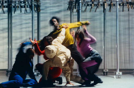 A dynamic group of five dancers wearing vibrant outfits ranging from yellow to pink strike expressive poses on a stage. Their movements are fluid, with one dancer reaching upwards, while others are bent or leaning, creating a sense of motion and collaboration. The background features metallic structures, suggesting a modern, industrial setting that contrasts with their lively performance.