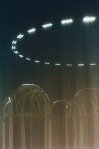 Soft lighting casts a mysterious yet serene atmosphere over the scene. Glowing arches rise towards a hazy sky, partially shrouded by semi-transparent fabric, creating an ethereal effect. A circular arrangement of light trails draws attention, hovering above the arches, suggesting motion or an artistic light installation. The composition evokes curiosity and a sense of wonder.