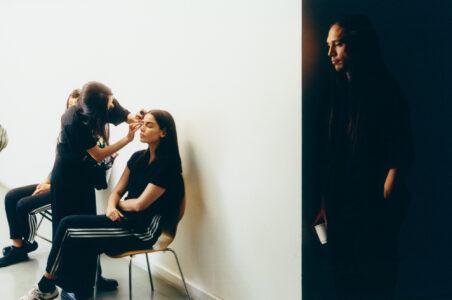 A makeup artist carefully applies makeup to a seated person's face, who is wearing a black outfit with striped pants. Another person sits nearby in similar attire. A third person with long hair stands against a shadowed wall, holding a cup, with hands in pockets. The scene suggests preparation before an event, with a contrast between light and shadow.