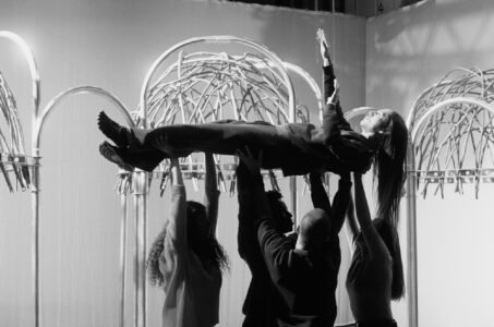 A monochrome scene shows a person being smoothly lifted horizontally by four individuals against an architectural backdrop. The group works together, supporting the person's limbs and torso with care and precision. The setting features curved, metallic structures resembling arches, contributing to an artistic and collaborative atmosphere. Shadows and lighting enhance the dramatic and focused mood of the moment.