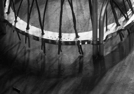 Shadows stretch across a textured surface, cast by metallic and organic elements. Thin rods arc from a smooth, light-colored band, with prominent shadows creating an intricate pattern. A smooth cylindrical column supports the structure, contrasting with rough, uneven shadows on the ground. The composition conveys a juxtaposition of industrial and natural forms in monochrome tones.