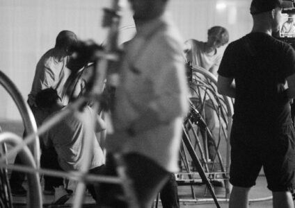 A group of people engage in what appears to be a film or photography setup, surrounded by various equipment. Some individuals are adjusting devices, while others stand observing. The scene is captured in black and white, creating a vintage, cinematic ambiance. Soft lighting and shadows emphasize the collaborative atmosphere of the production environment.
