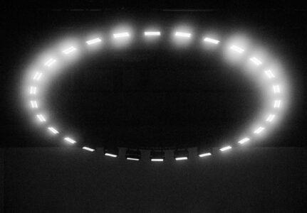 In a dark environment, an illuminated circular arrangement of bright, evenly spaced rectangular lights forms a gentle arc. The surrounding area is shrouded in shadows, enhancing the luminosity of the lights. The pattern suggests a futuristic or technological atmosphere, with a sense of mystery and intrigue, as if observing an installation or object from science fiction.