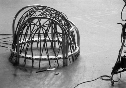 A black-and-white scene shows a complex structure of intertwining metal and cable-like components forming a dome shape. It rests on a plain surface, with loose wires scattered around. Nearby, a wheeled piece of equipment with circular elements and a handle is visible. Shadows and various objects create an industrial or workshop-like atmosphere.