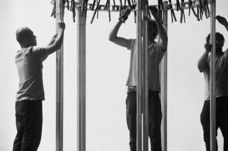 Three individuals are working together on a vertical structure, adjusting components near the top. The scene appears collaborative, with one person closely observing and the others actively manipulating the assembly with tools. The background is plain, directing focus to the figures and the intricate construction. This monochrome setting emphasizes teamwork and attention to detail in their task.