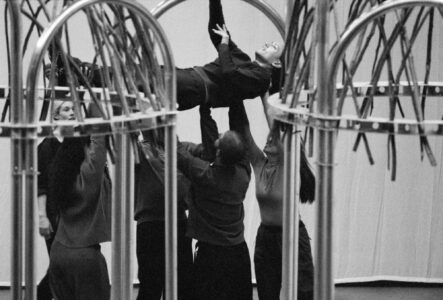 A group of people is collaboratively holding a person aloft in what seems to be a creative performance or rehearsal. The setting features tall, curved metal structures that frame the scene. The individuals are dressed in dark garments, and the atmosphere is focused, conveying a sense of teamwork and expressive movement in a monochrome palette.