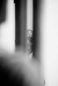 A monochrome scene captures the partial view of an individual’s face through vertical barriers, creating a mysterious and dramatic composition. The play of light and shadows adds depth and intensity, while the blurred elements enhance the enigmatic atmosphere. The focus is on the person’s eye and forehead, partially obscured, suggesting intrigue and contemplation.
