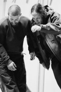 Two individuals appear engaged in a focused activity, both leaning forward with intent expressions. One wears a ribbed sweater, while the other sports a hooded jacket. Their posture suggests concentration or collaboration. Captured in black and white, the scene exudes a sense of movement and communication amid an undefined background, emphasizing their interaction and shared purpose.