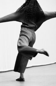A person with curly hair is captured mid-movement, wearing a striped, loose-fitting outfit that includes a long-sleeved top and wide pants. The individual appears to be spinning or dancing, with one foot raised and the other touching the ground. The background is a plain, light-colored wall, emphasizing the dynamic motion and flow of the scene.