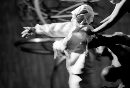 A dynamic, blurred scene of motion captures several figures in abstract movement. One wears a white garment, creating a sense of theatricality and drama. Arms are extended, suggesting a coordinated dance or performance. The composition is monochromatic with high contrast, emphasizing movement and expression. The background features indistinct, circular shapes, adding depth and intrigue to the scene.
