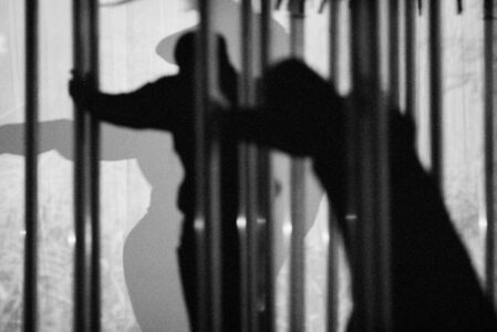 A monochromatic scene features silhouettes against a textured backdrop, evoking a sense of mystery. The figures appear dynamic, engaged in motion or interaction. Strong contrasts and blurred vertical lines create an abstract, dreamlike quality. One silhouette wears a hat, adding intrigue and balance to the composition. The overall atmosphere suggests shadow play and artistic expression.
